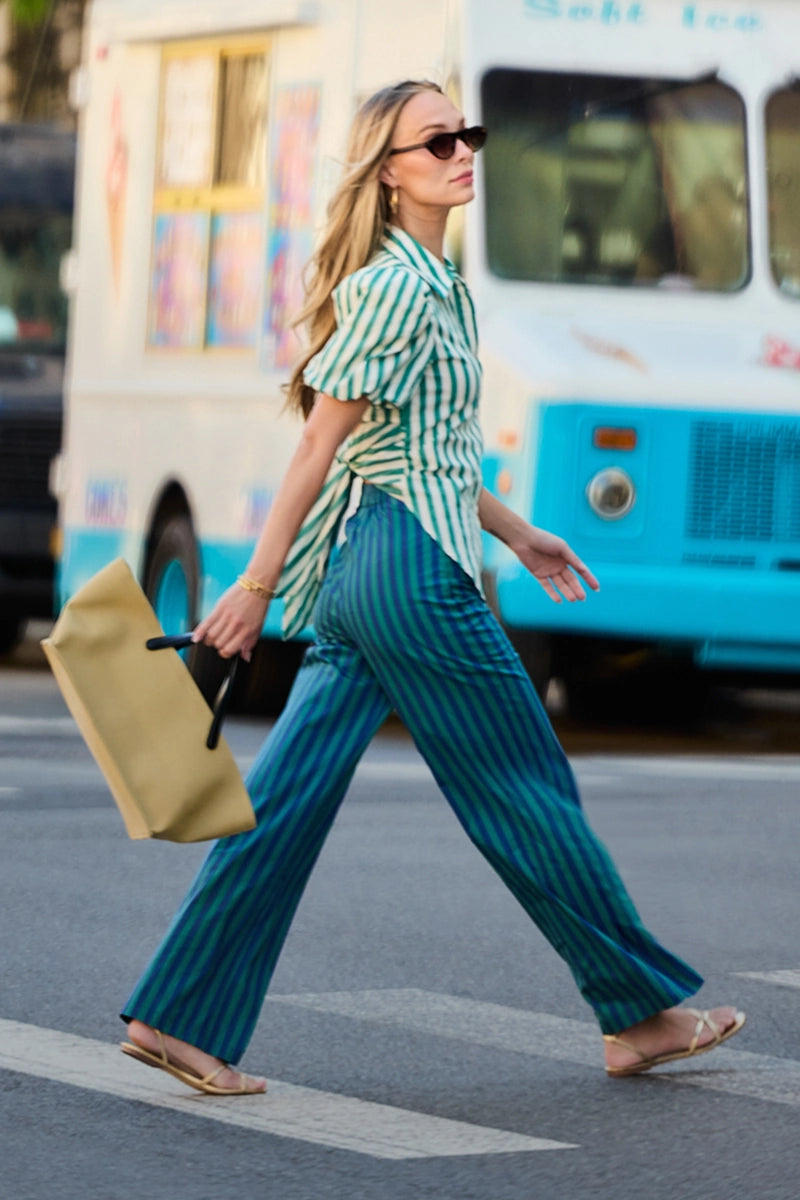 A woman walking across a street crosswalk wearing a green and white striped short-sleeve button-up shirt and matching teal and navy vertically striped wide-leg trousers, carrying a tan paper bag. mobile