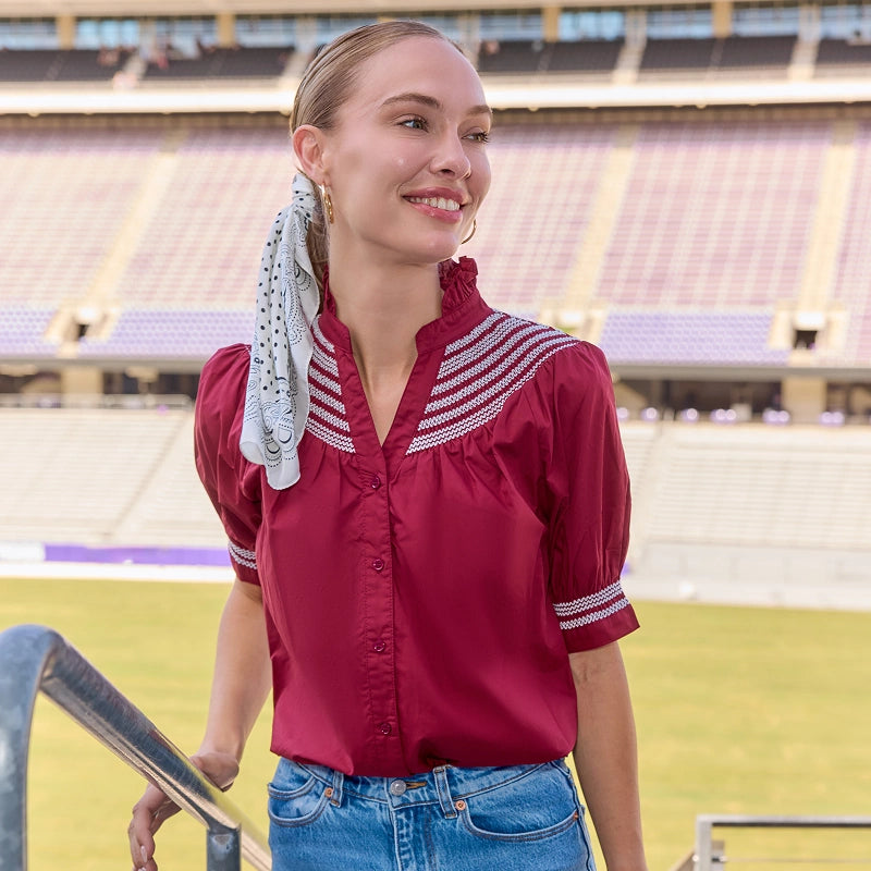 A woman wearing a burgundy short-sleeve blouse with white striped yoke detailing and blue jeans, standing in a large outdoor stadium with empty purple seating in the background.