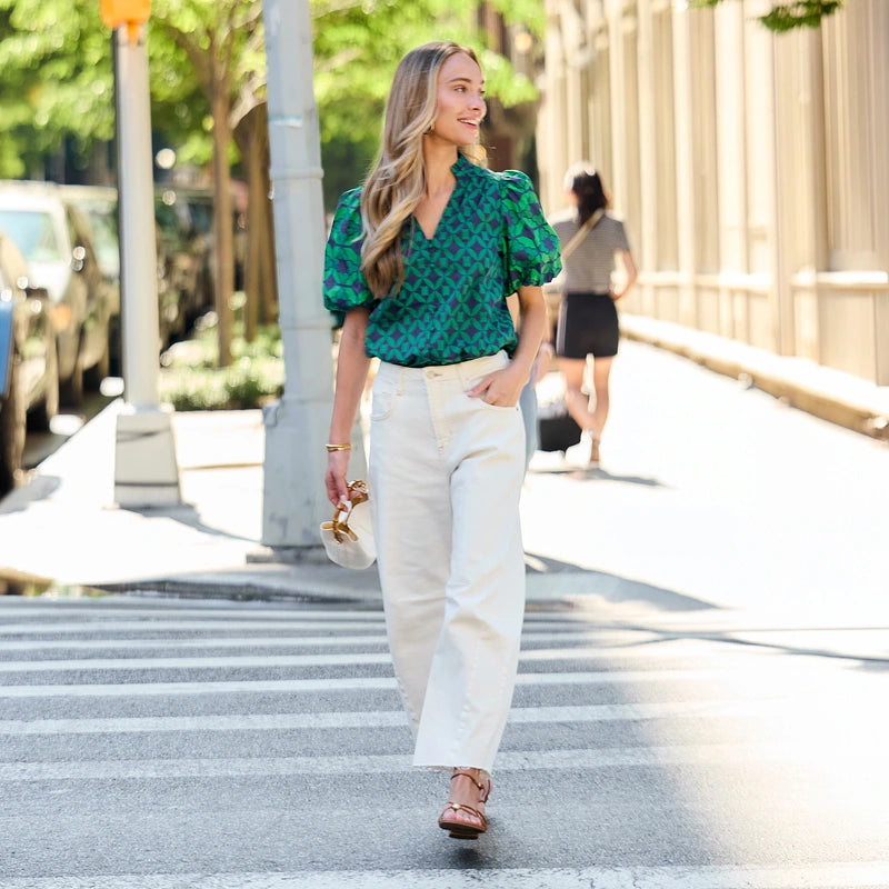 A woman walking on a city crosswalk wearing a green patterned puff-sleeve blouse, white wide-leg trousers, and brown sandals, holding a small beige handbag. collection