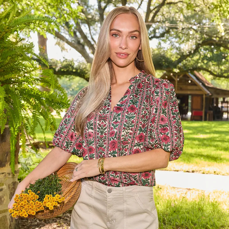 A woman stands outdoors in a garden setting, wearing a short-sleeve button-up blouse with a dense floral and botanical print in red, green, and beige tones, paired with light beige pants. She is holding a small wicker basket filled with yellow flowers.