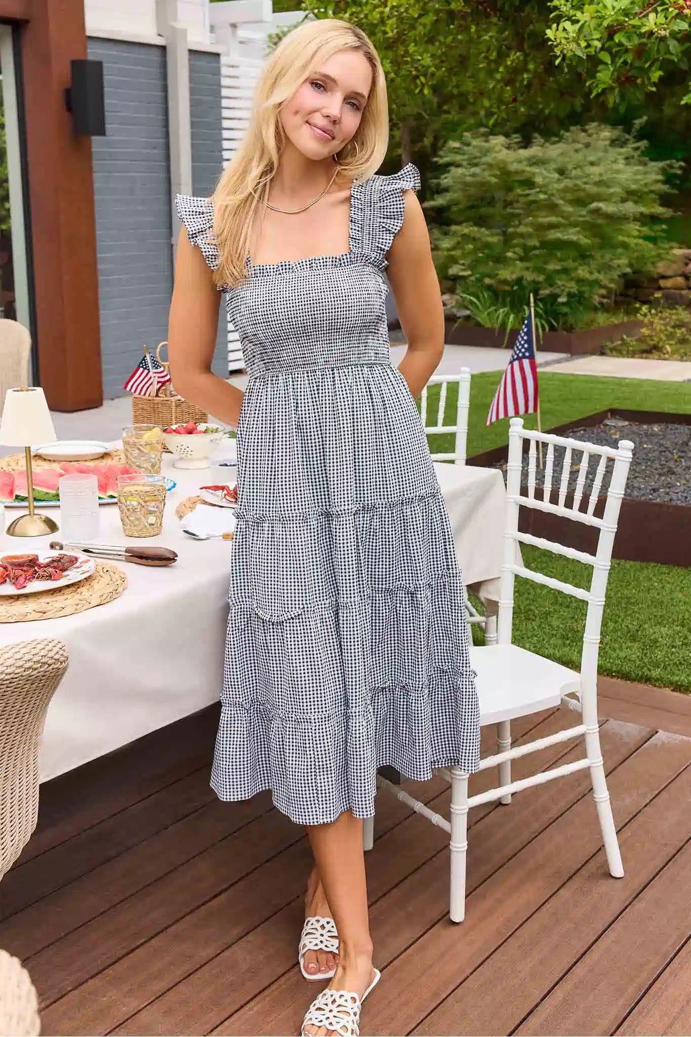 Woman in a blue checkered dress standing on a wooden deck with a table and chairs in the background.