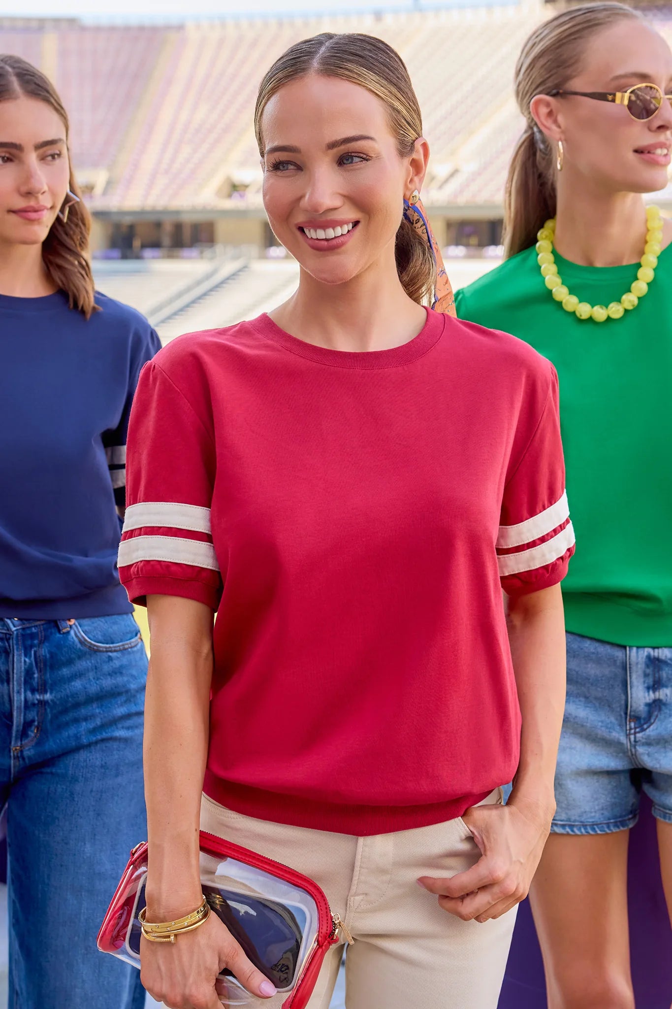 A woman in a bright pink short-sleeve crewneck sweatshirt with white stripes on the sleeves, paired with beige pants, standing in an outdoor stadium setting with other women in the background.