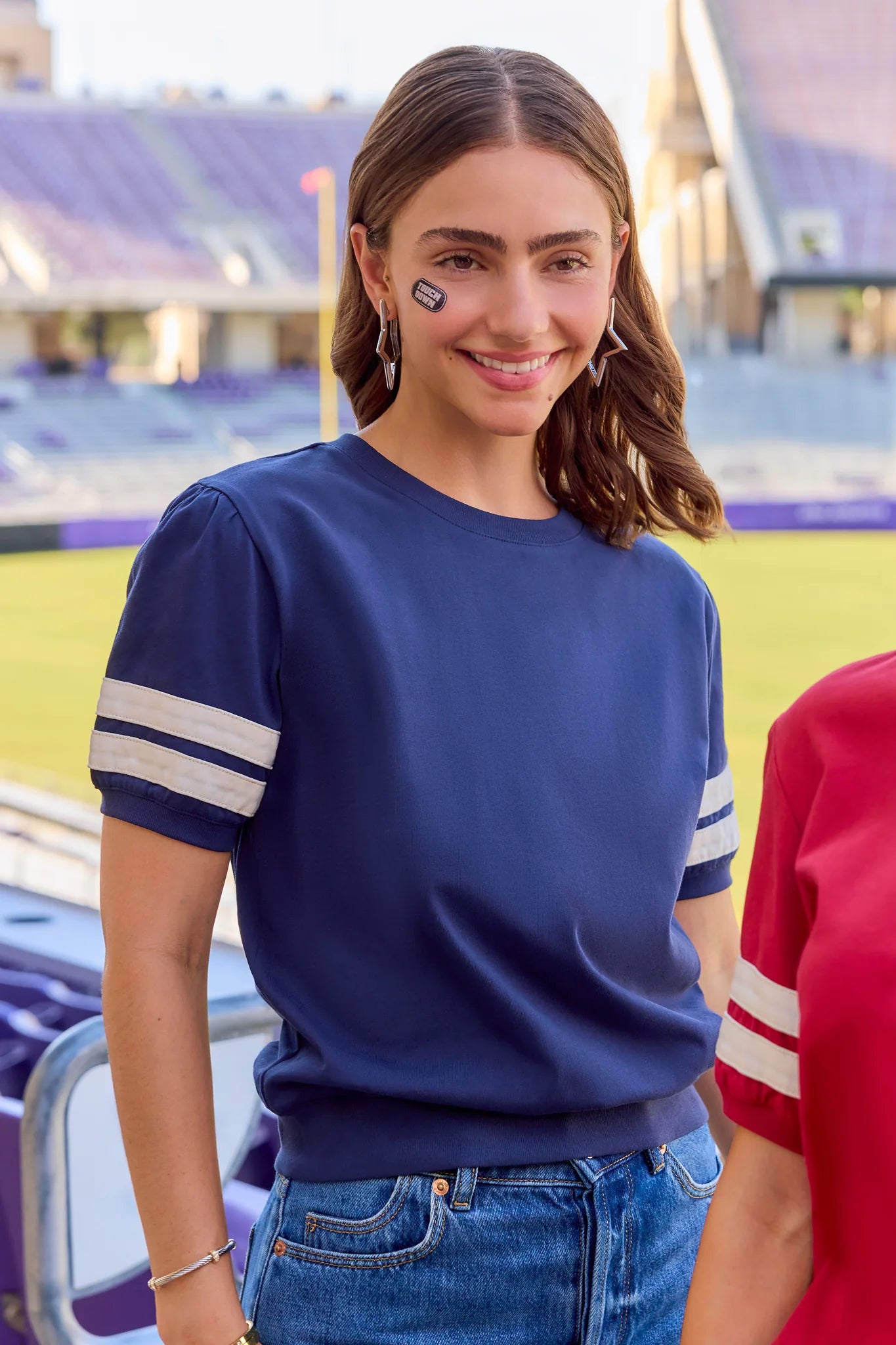 A woman wearing a navy blue short-sleeve t-shirt with white stripes on the sleeves, standing in a stadium setting.