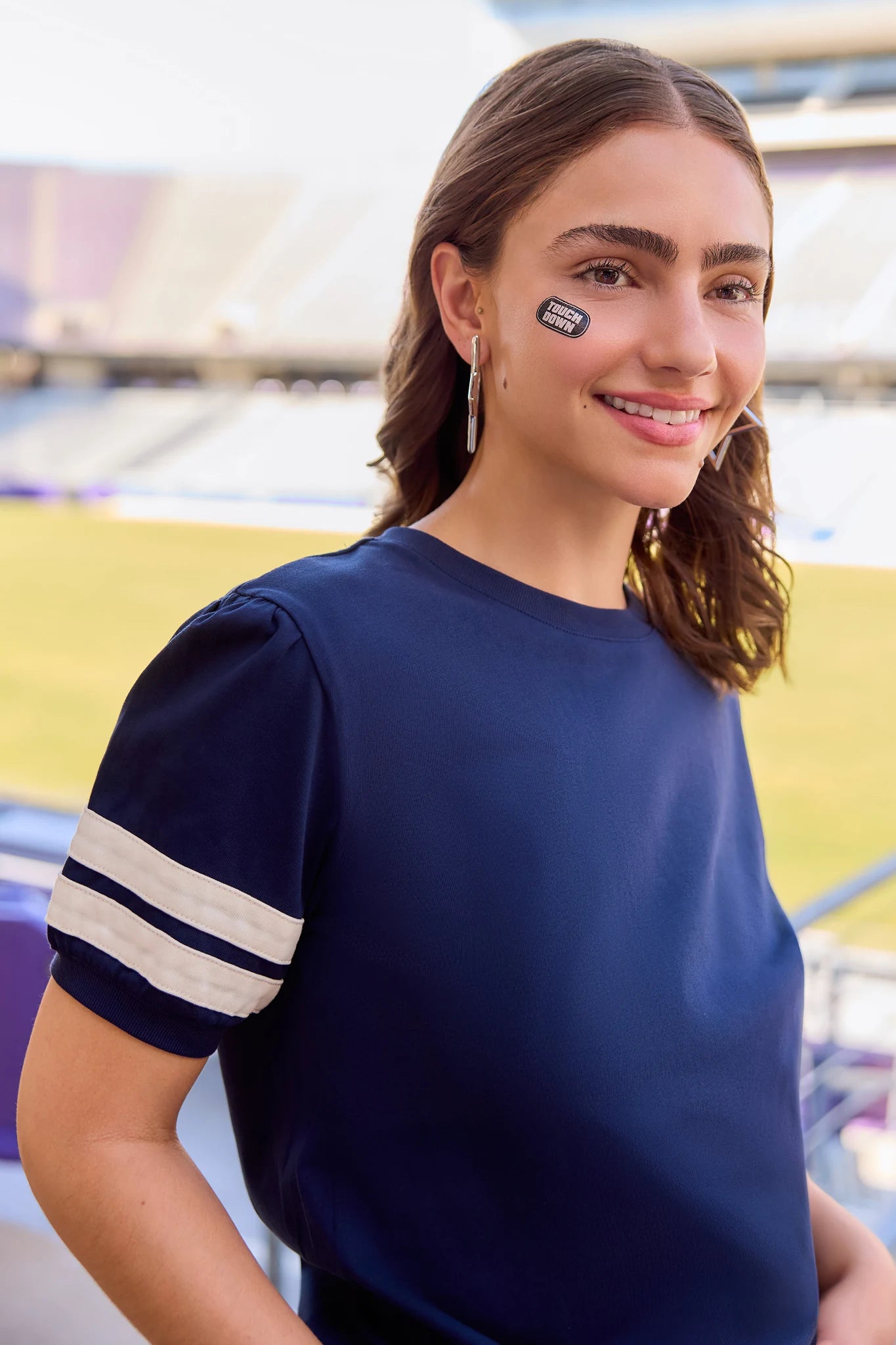 A woman wearing a navy blue short-sleeve t-shirt with white stripes on the sleeves, standing in a stadium setting.