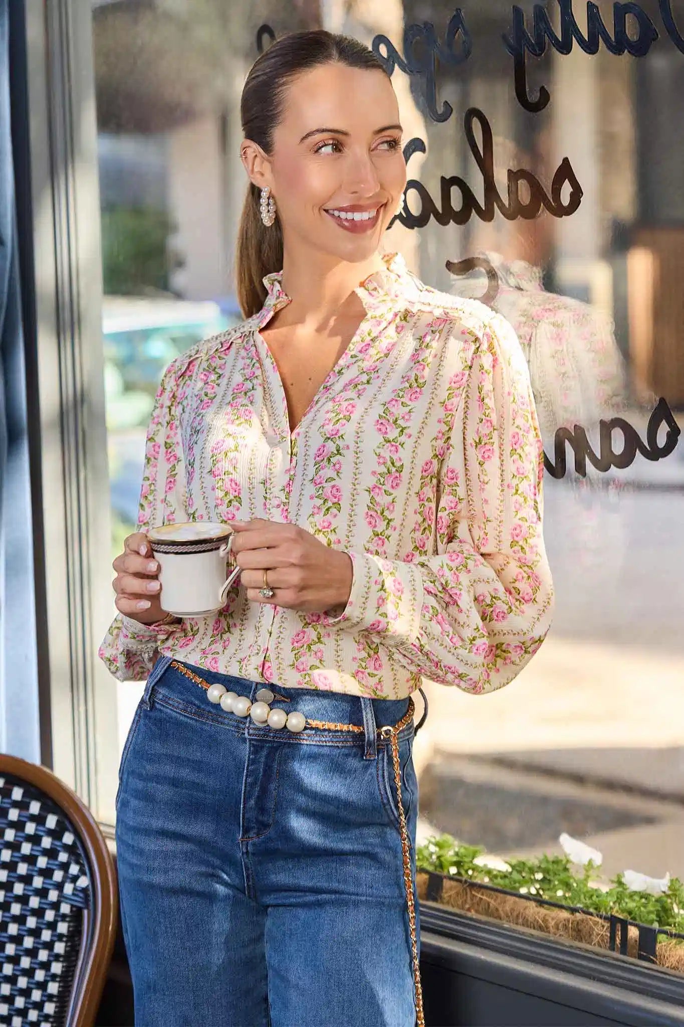 Woman in a floral blouse and blue jeans holding a coffee cup, standing in front of a store window.