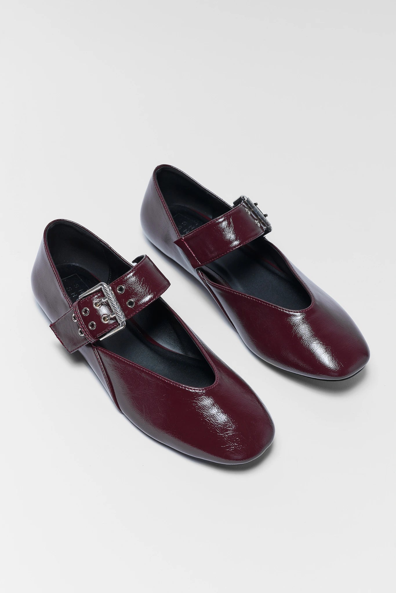 A pair of women's flats in glossy burgundy faux leather, featuring a single strap with a silver-tone buckle and eyelets, displayed on a light gray background.