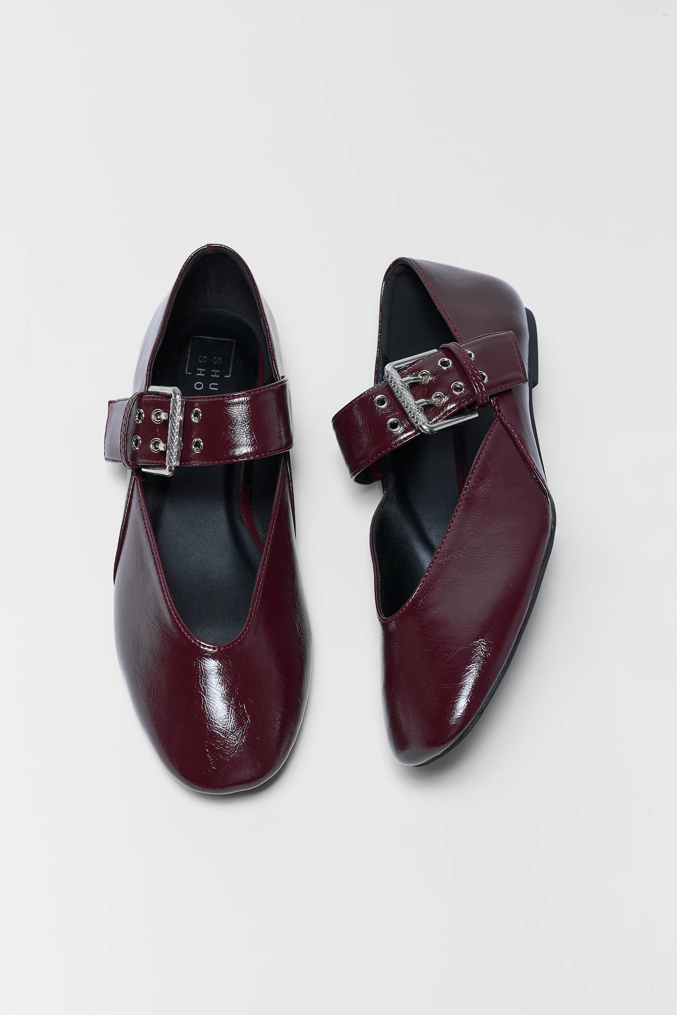 A pair of women's Mary Jane flats in glossy burgundy leather, featuring a single wide strap with a silver-tone buckle and metal eyelets, shown on a light gray background.