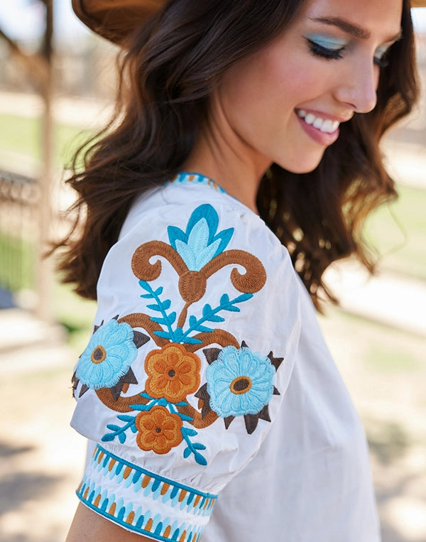 Woman wearing a white Eileen top with blue and brown floral embroidery outdoors.