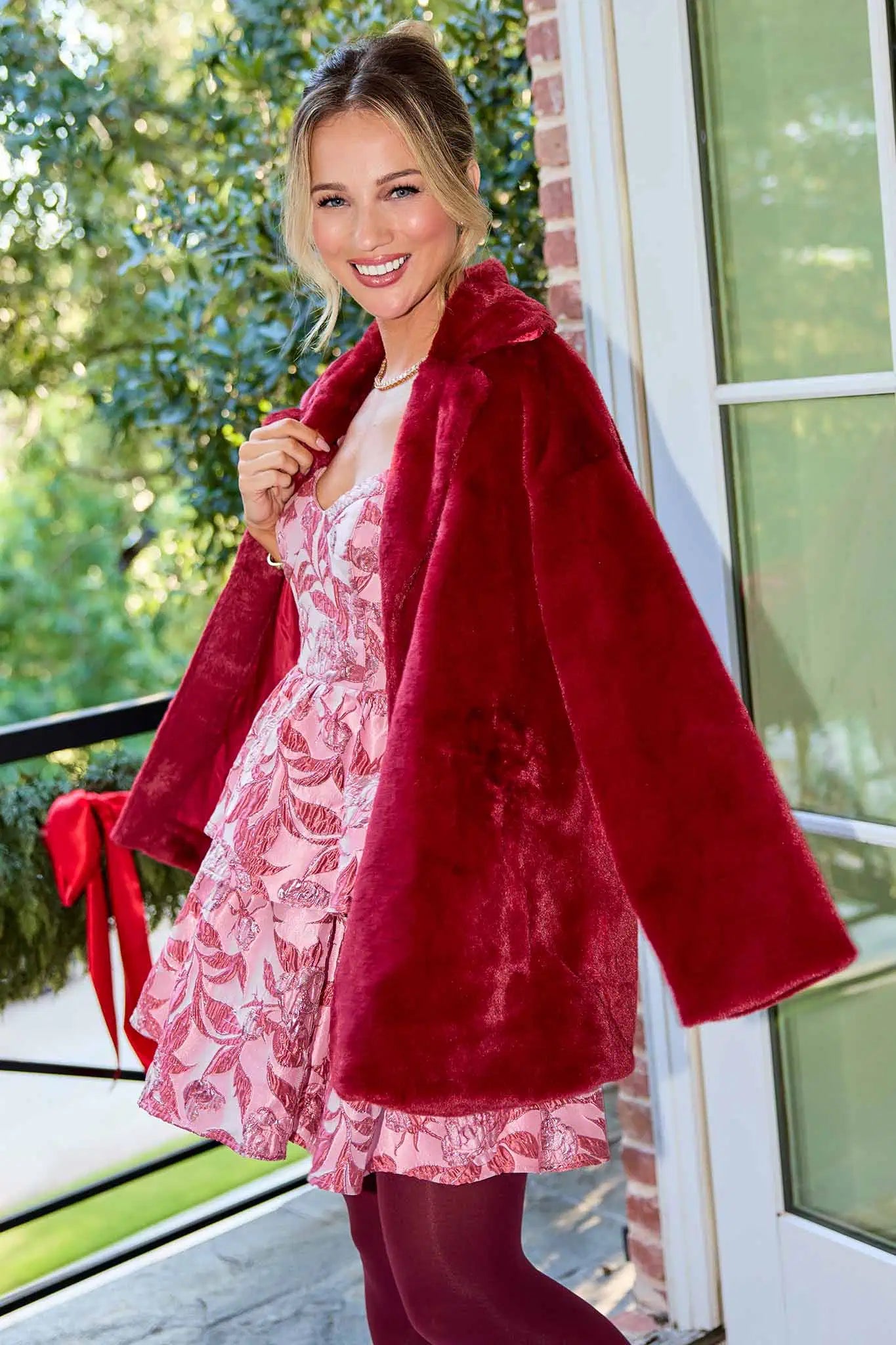 Woman wearing a red coat over a pink floral dress on a balcony.