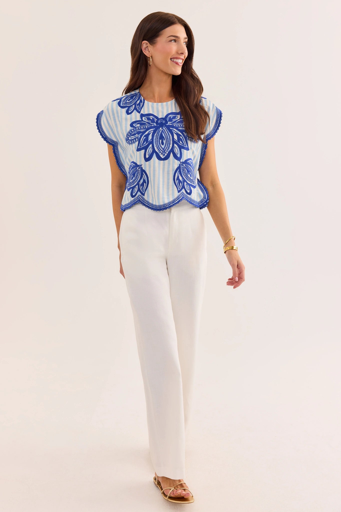 Woman wearing a blue embroidered top and white pants on a light background