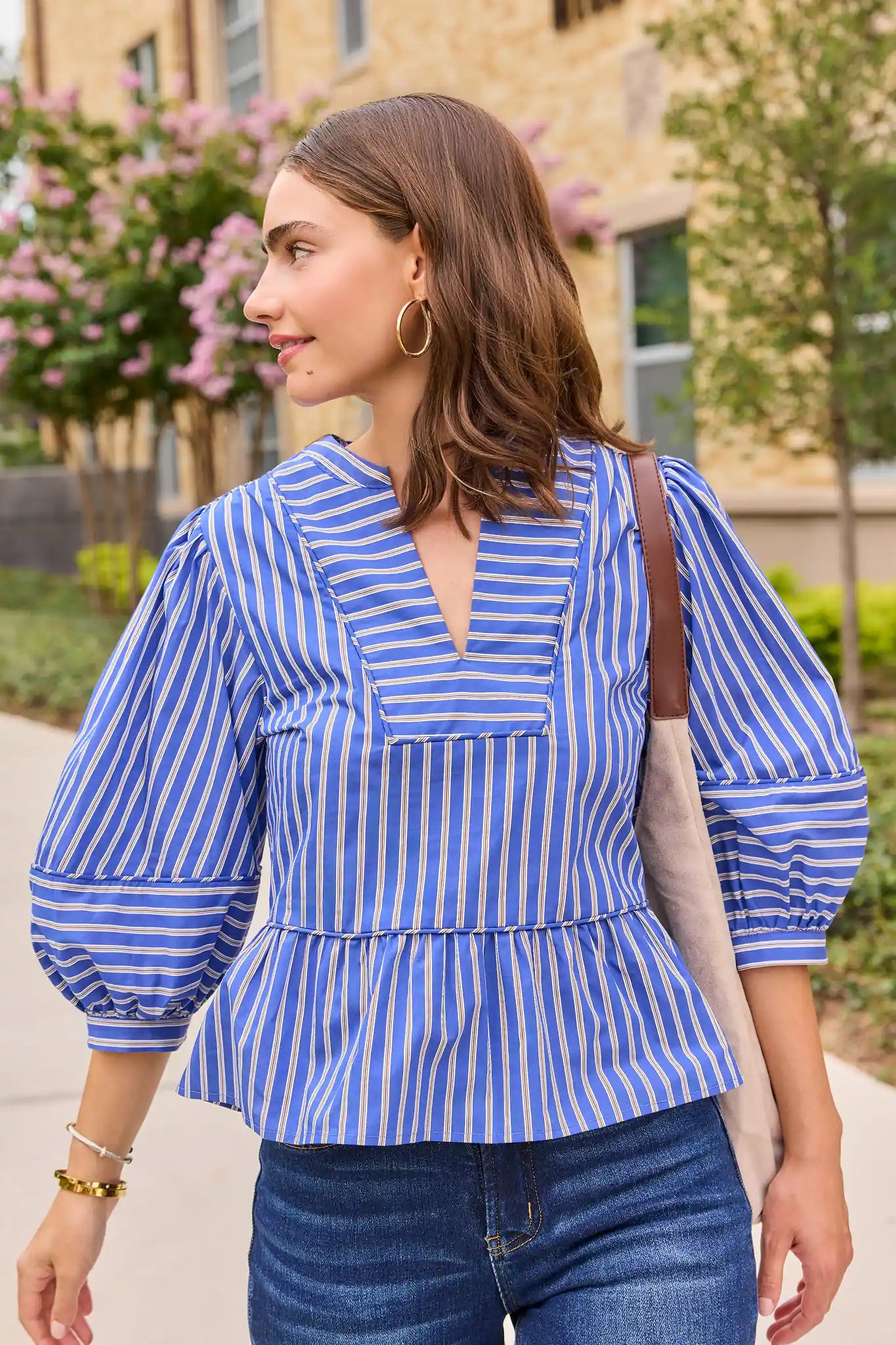 A woman wearing a blue and white vertically striped blouse with puff sleeves and a peplum hem, paired with blue jeans and a beige shoulder bag, standing outdoors in front of a building and trees.