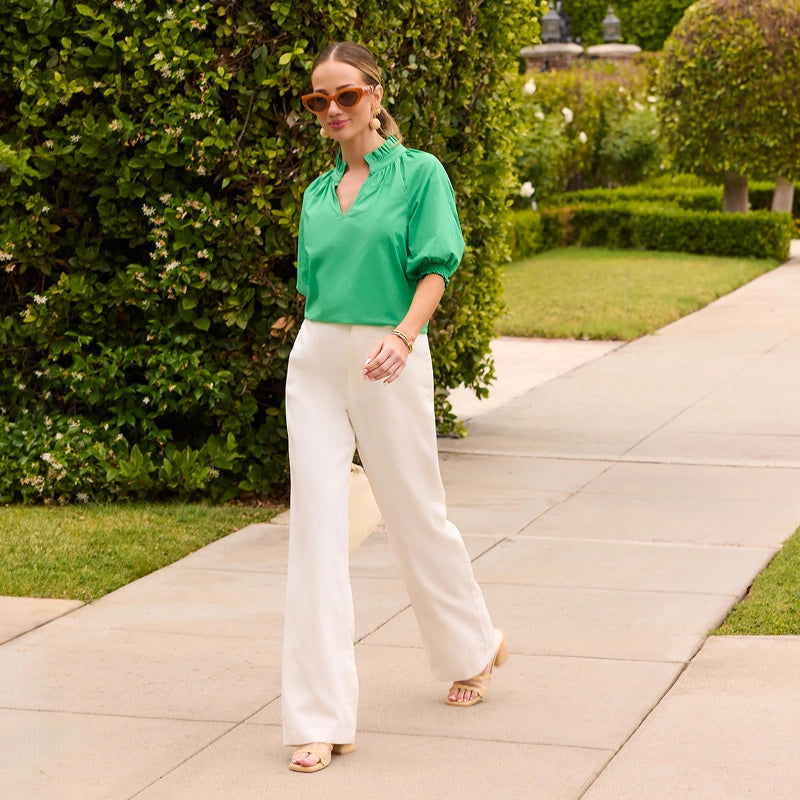 Woman in a green blouse and white pants standing on a sidewalk with greenery in the background collection