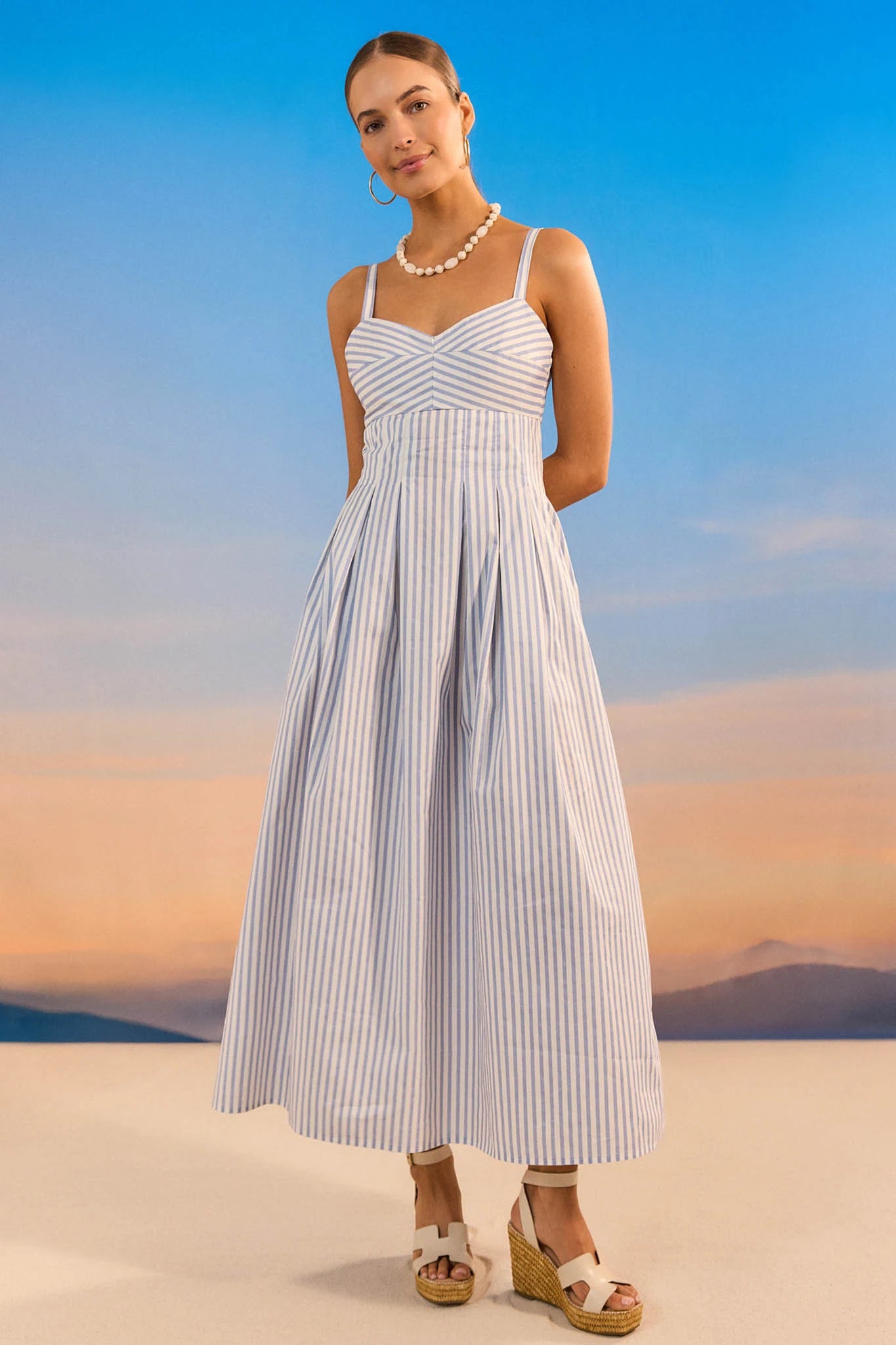 Woman in a white and blue striped dress standing against a sunset sky.