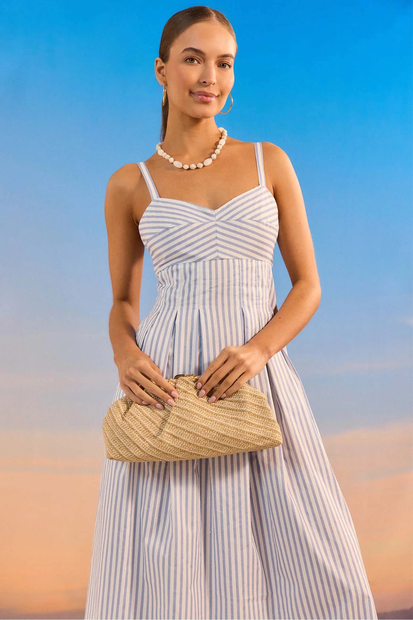 Woman in a striped dress holding a straw clutch against a blue sky and sand background