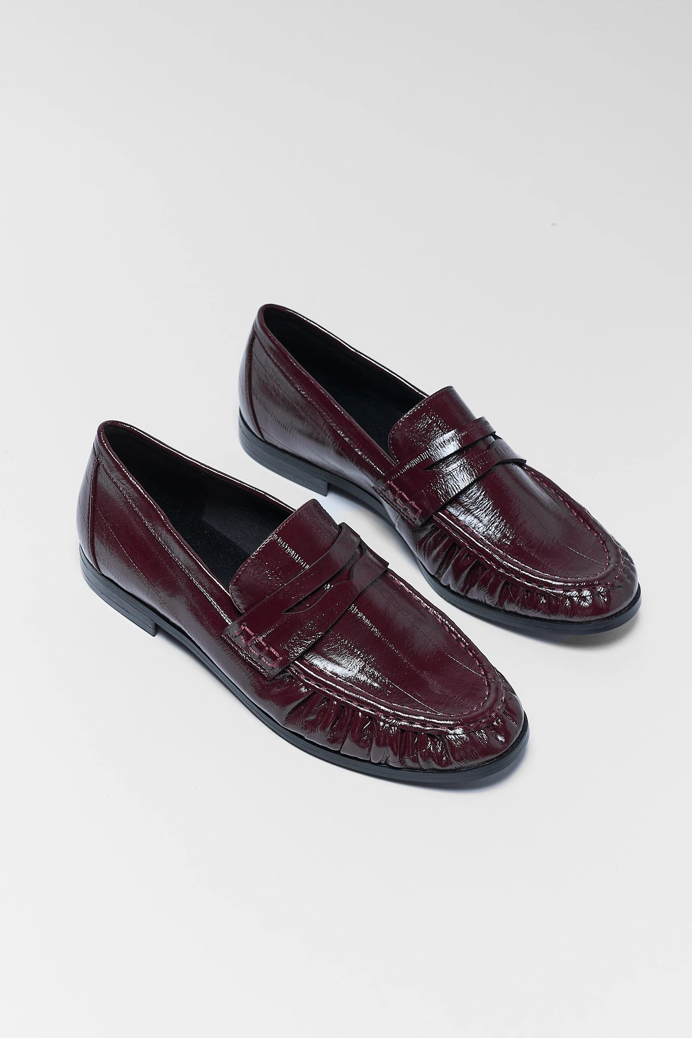 A pair of glossy burgundy leather penny loafers with a textured crocodile-embossed pattern, displayed on a light gray background.