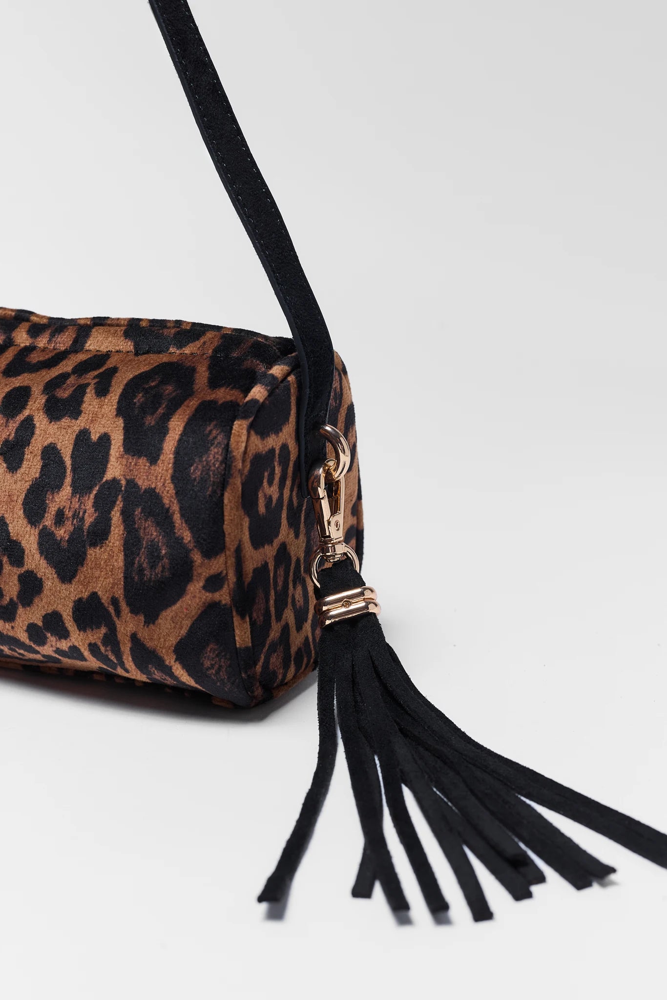 Close-up of a leopard-print handbag with a black strap and a black tassel charm featuring gold-tone hardware, on a light gray background.
