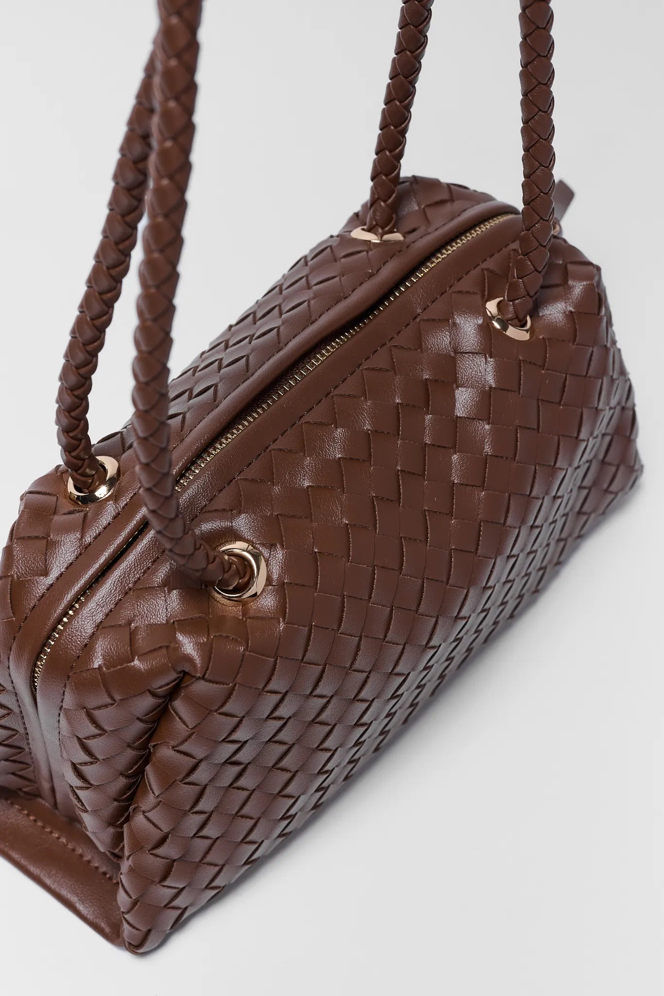A brown woven leather handbag with braided shoulder straps and gold-tone hardware, photographed on a clean light gray background.