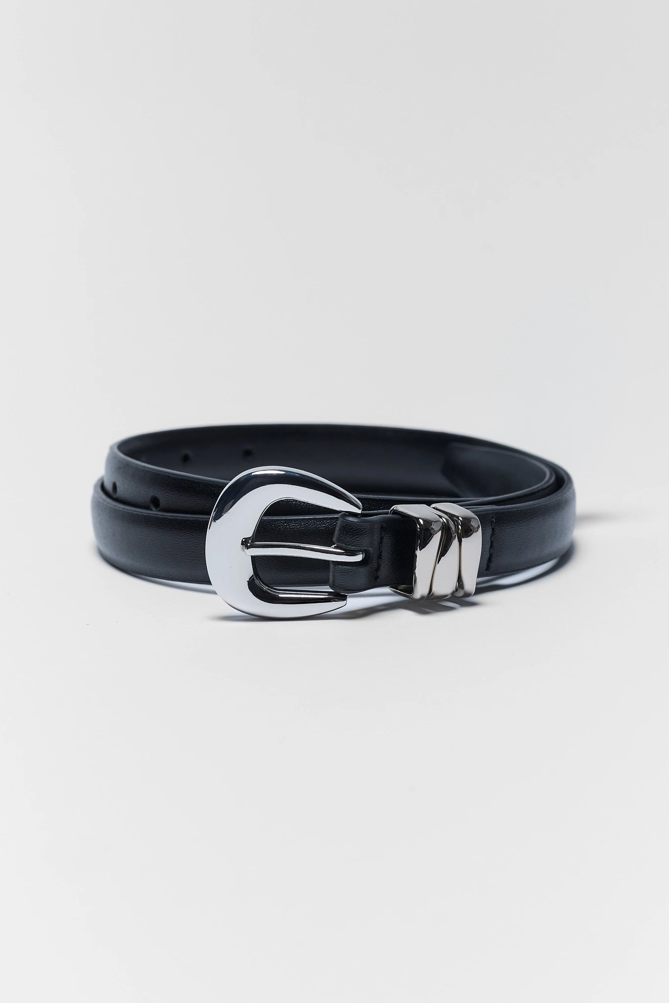 A black leather belt with a polished silver-tone metal buckle, coiled on a light gray studio background.