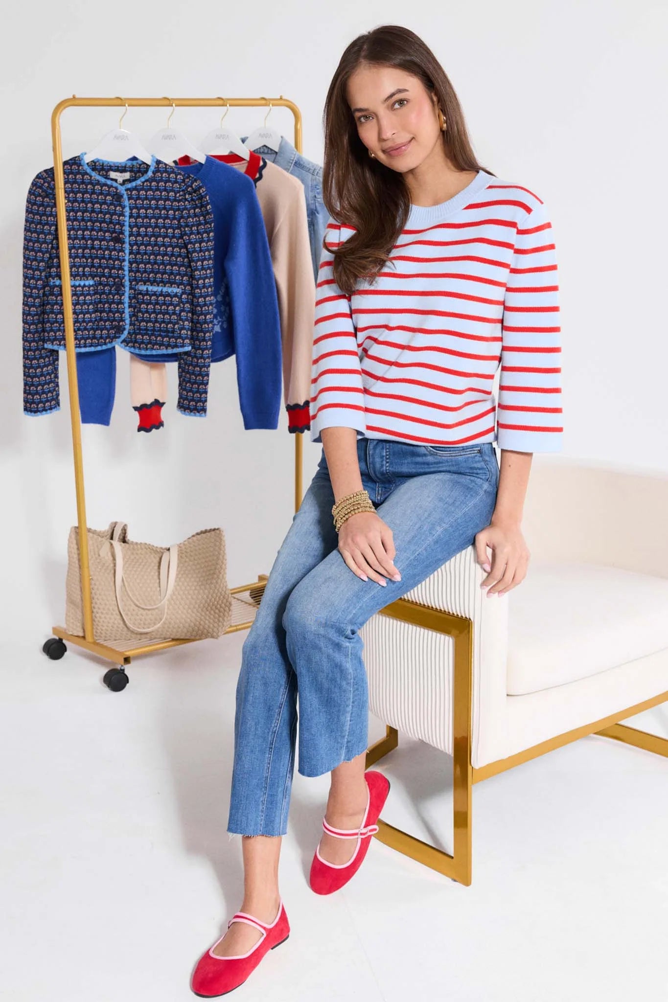 Woman sitting on a chair wearing a striped shirt and jeans, with a clothing rack in the background.