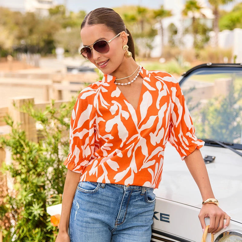 Woman in an orange and white patterned blouse and blue jeans standing next to a white car. collection