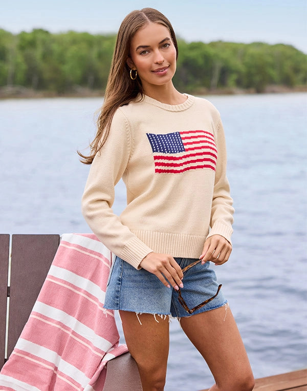 Woman wearing a beige sweater with an American flag design by a lake. navigation