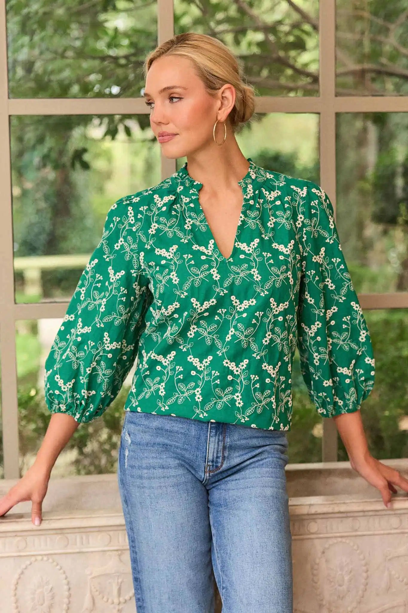 Woman wearing a green floral blouse and blue jeans standing in front of a window with a view of trees.