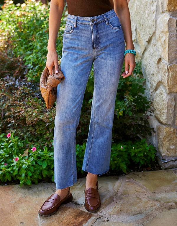 Women wearing blue Kelsey jeans and brown Hardi loafers standing outdoors with a stone wall and plants in the background