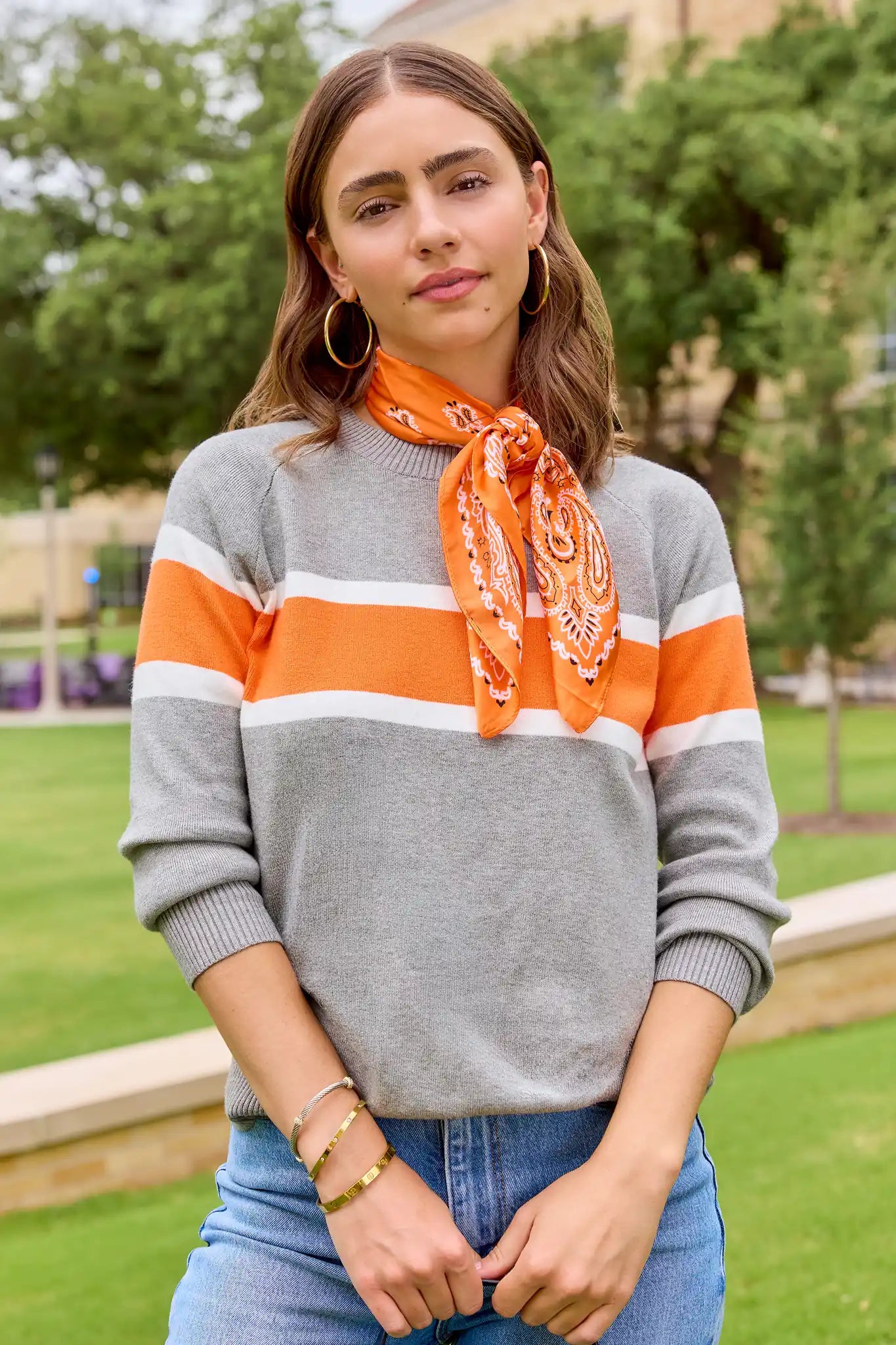 A woman wearing a gray knit sweater with orange and white horizontal stripes, styled with an orange paisley neck scarf and blue jeans, standing outdoors on a grassy lawn.