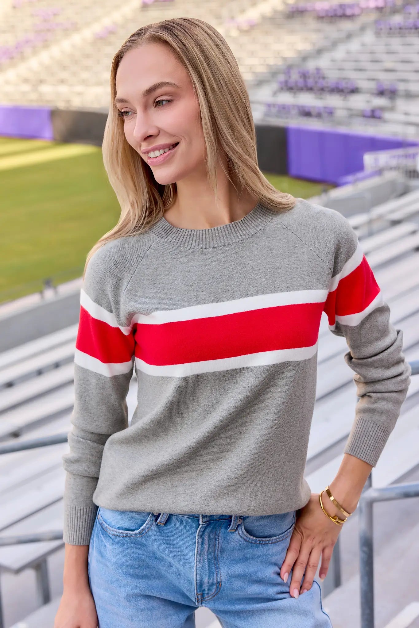 A woman wearing a heather gray crewneck sweater with bold horizontal red and white stripes across the chest and shoulders, paired with blue jeans, standing in an outdoor stadium setting.
