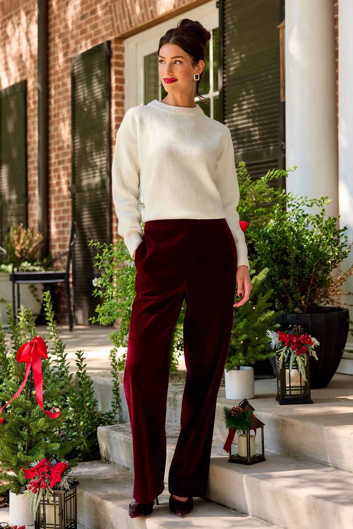 Woman in a white sweater and Evie Cranberry Velvet Pants standing on steps with decorative plants and lanterns.