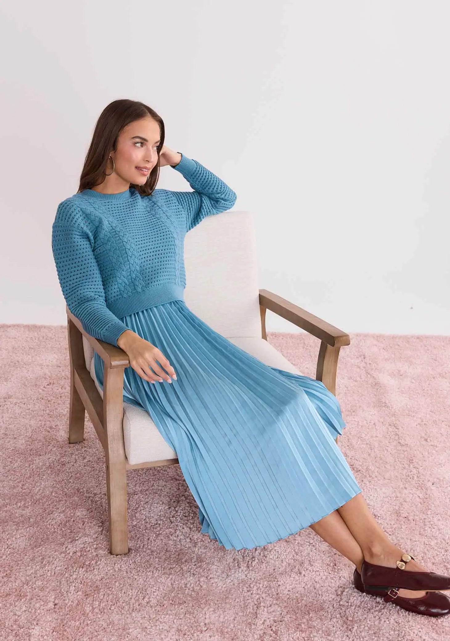 Woman wearing a Gabbie Dusty Blue Two Piece Midi Dress sitting on a wooden chair against a light pink background