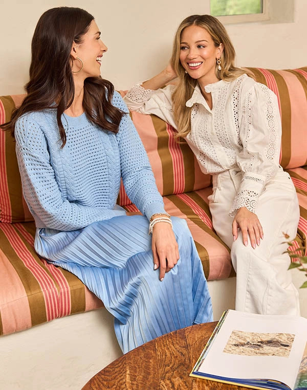 Two women sitting on a striped couch, one in a blue sweater and pleated skirt, the other in a white lace top and pants.