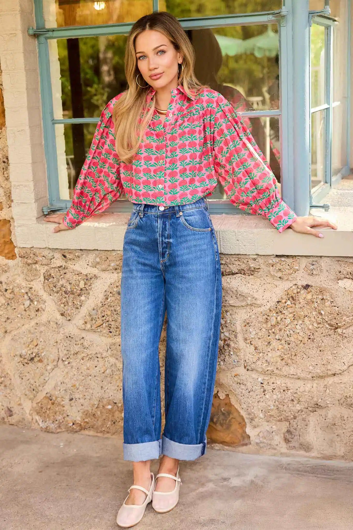 Woman wearing a red and white patterned blouse and blue jeans standing in front of a stone wall.