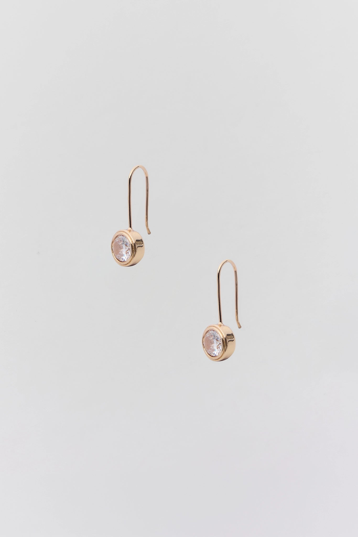 A pair of rose gold drop earrings with a small round clear gemstone in a bezel setting, suspended from simple curved hooks, shown on a light gray background.