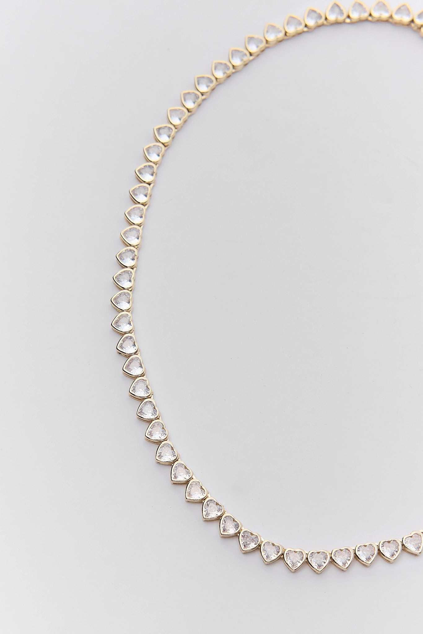 A gold tennis necklace featuring small, clear heart-shaped gemstones set in a gold chain, photographed on a light gray background.