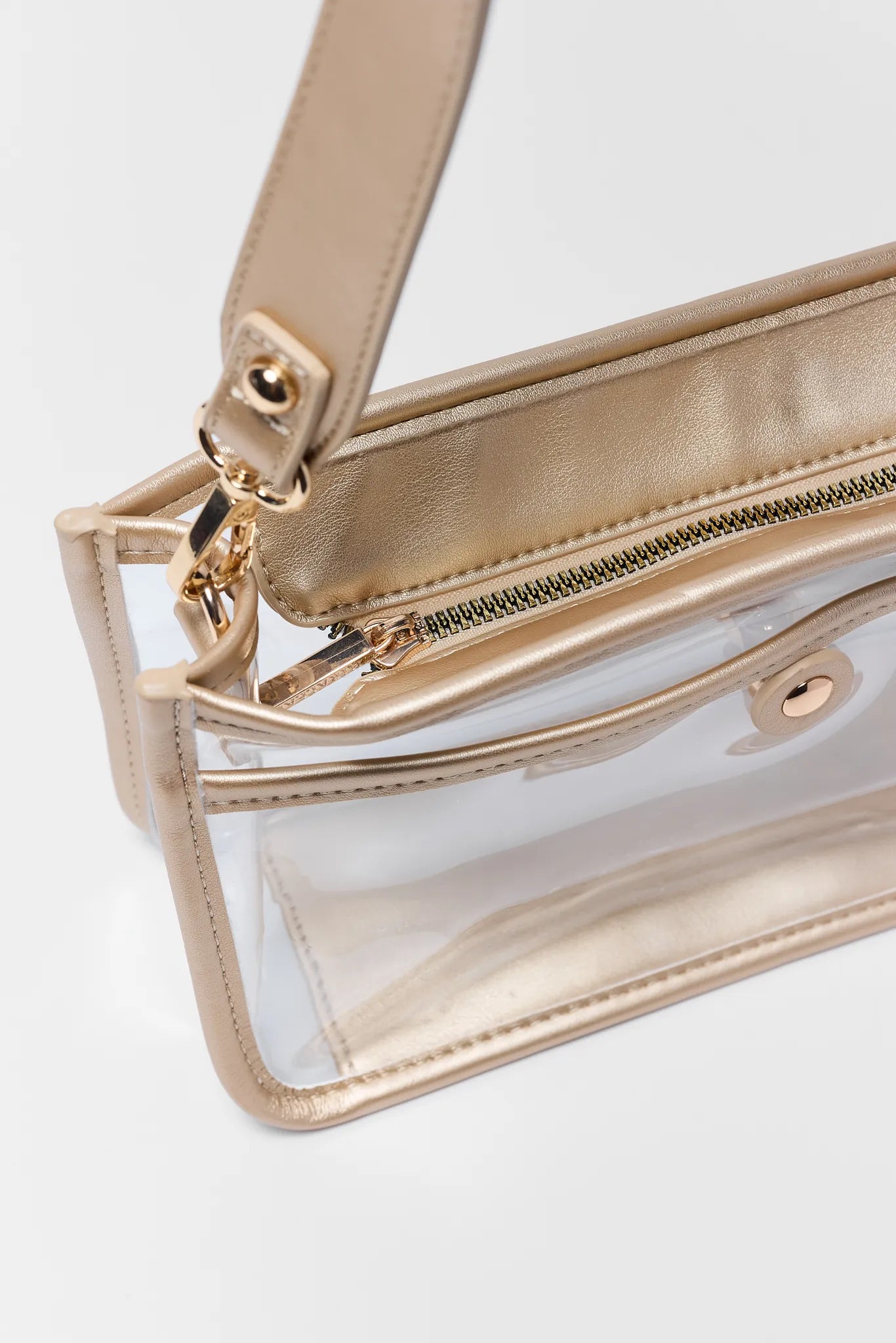 A close-up of a clear plastic pouch bag with metallic gold trim, zipper, and strap, shown on a light gray background.