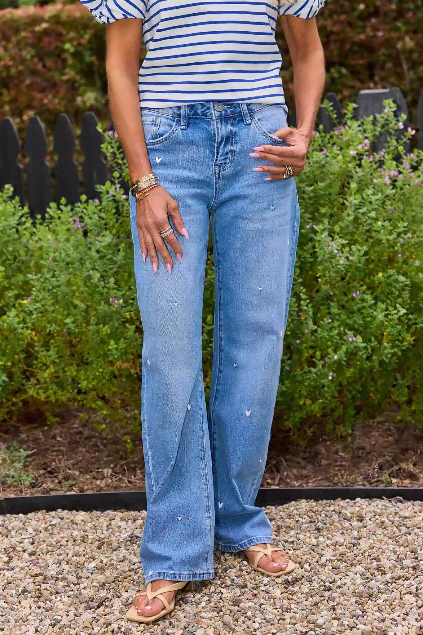 Person wearing blue jeans and a striped shirt standing outdoors with greenery in the background