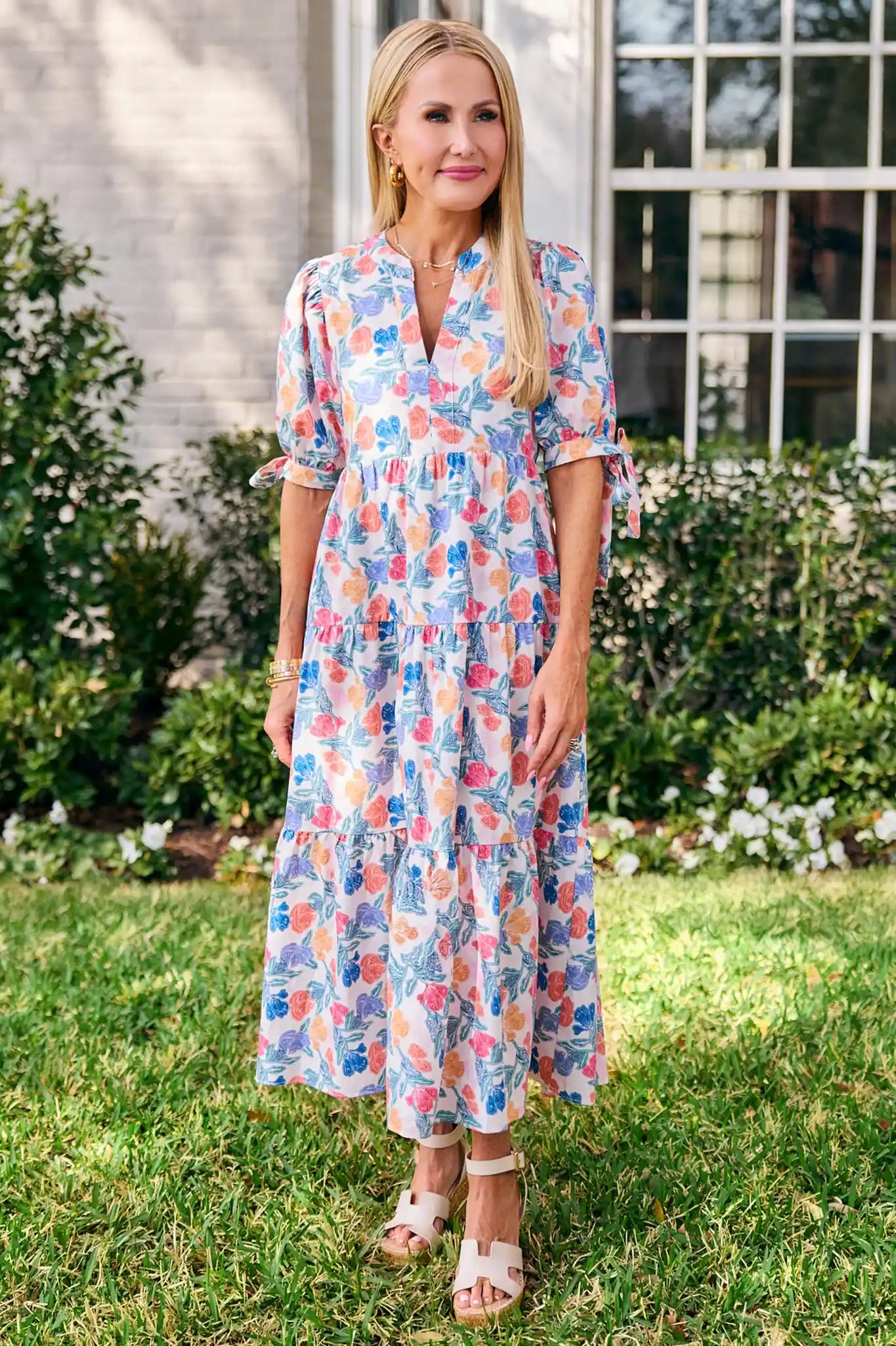 Woman in a floral dress standing outdoors with greenery and a building in the background