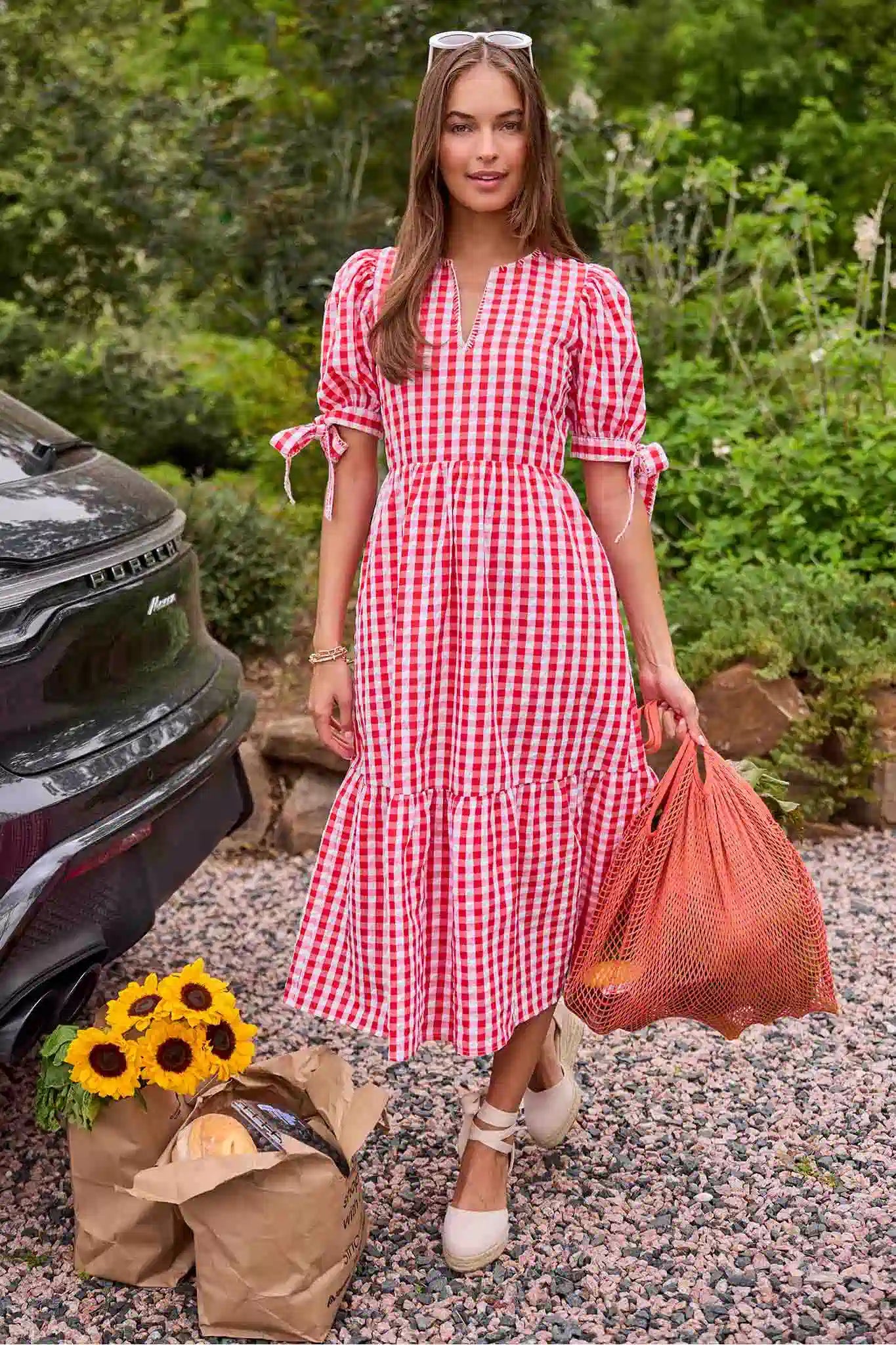 Woman in a red and white checkered dress standing outdoors with a car and sunflowers in the background.