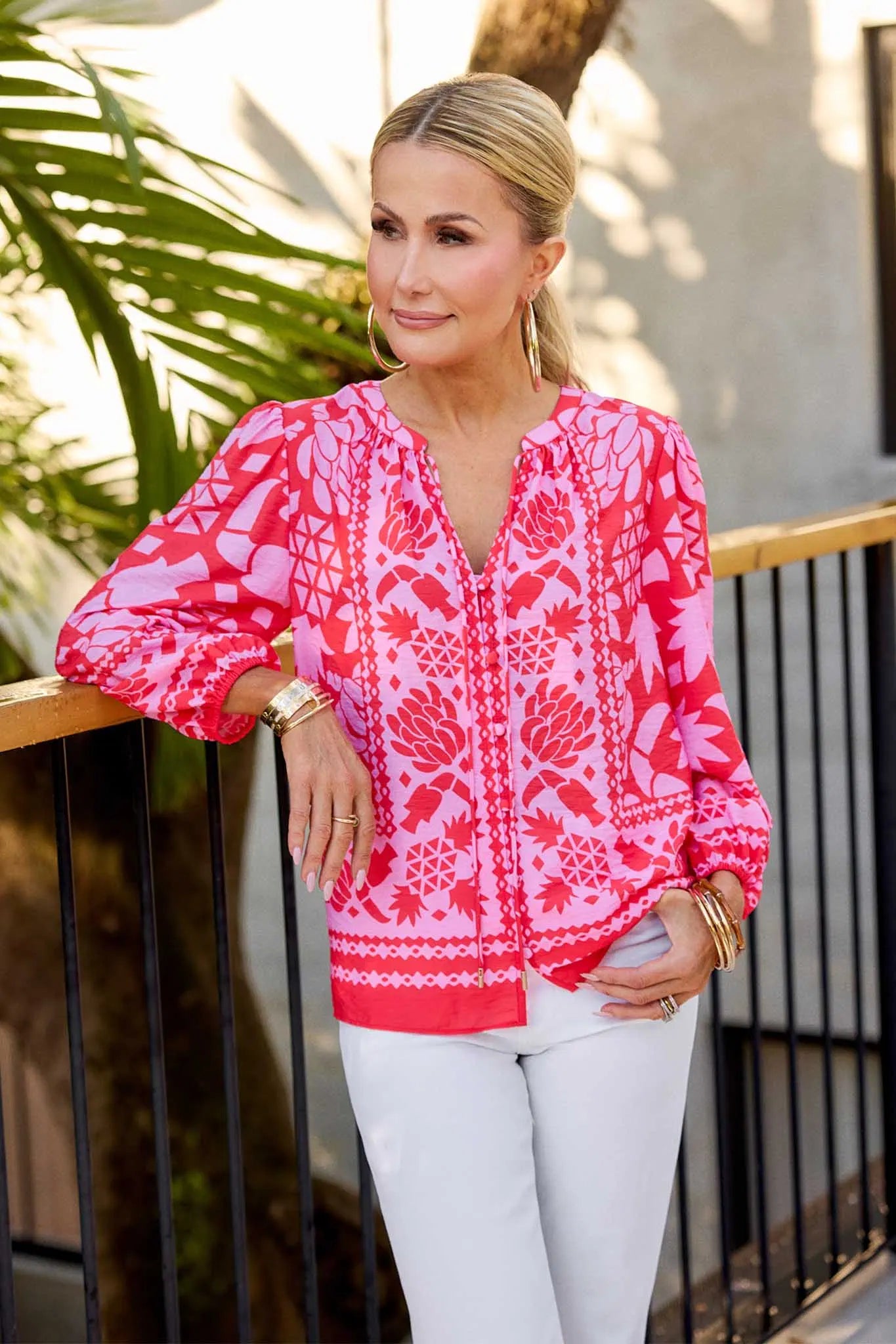 Woman wearing a pink patterned blouse with white pants standing on a balcony.