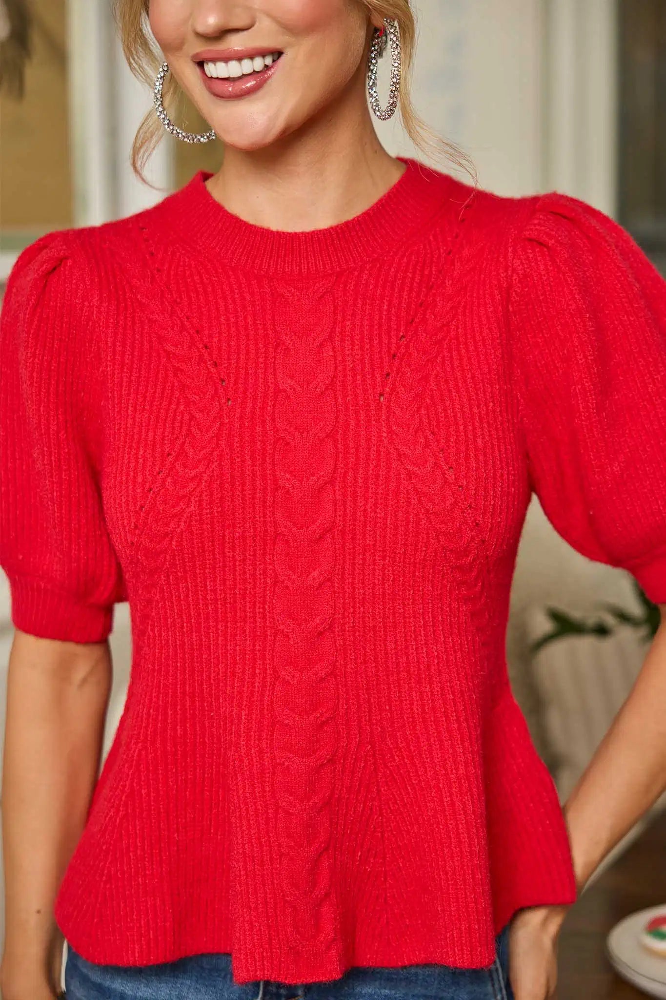 Woman in a red hillary top and blue jeans standing in a room with decor elements.
