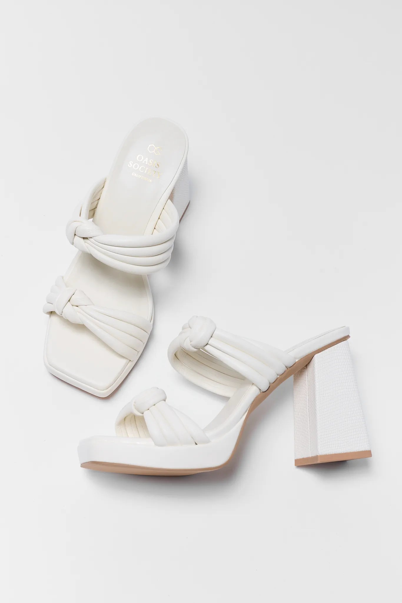 White high-heeled sandals with knot details on a light gray background