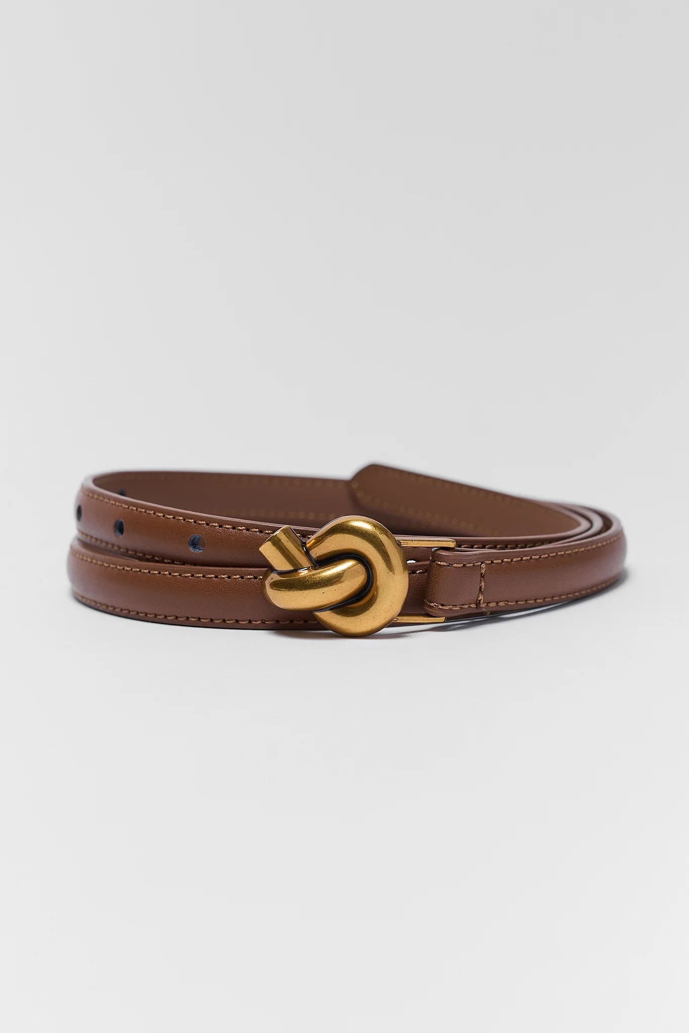 A brown leather belt with a distinctive gold-tone knot buckle, coiled on a light gray background.
