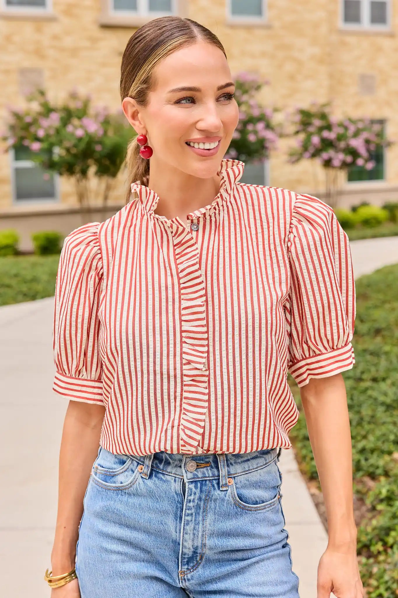 A woman wearing a red and white vertically striped short-sleeve blouse with ruffled trim and puff sleeves, paired with light blue jeans, standing outdoors in front of a beige brick building.