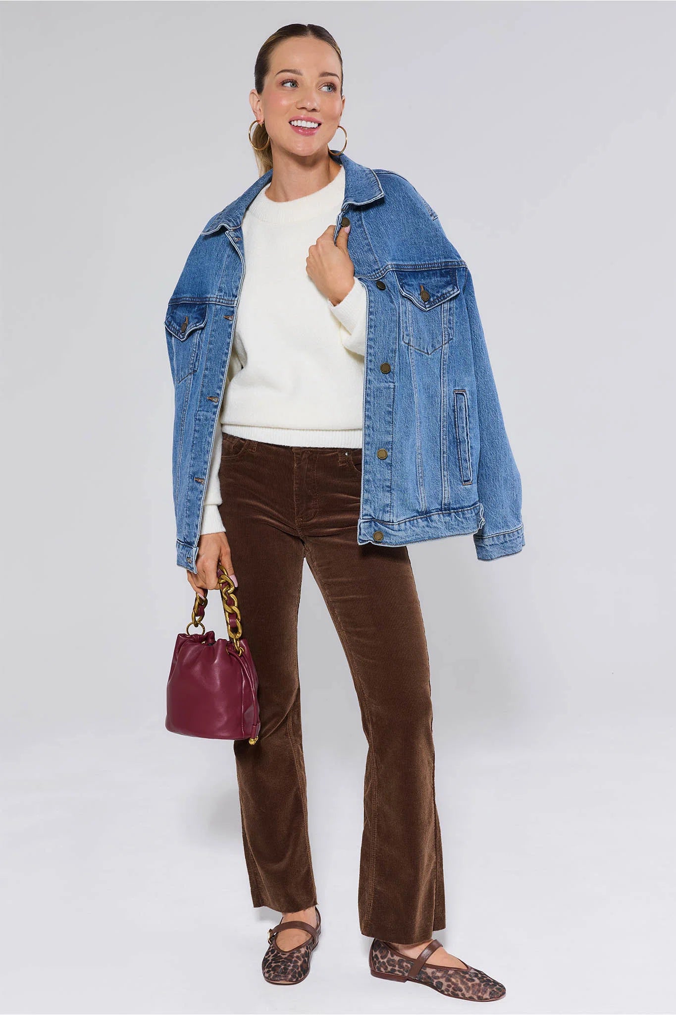 Woman wearing a blue denim jacket, white sweater, brown Kelsey Pants Corduroy, and carrying a red handbag on a white background
