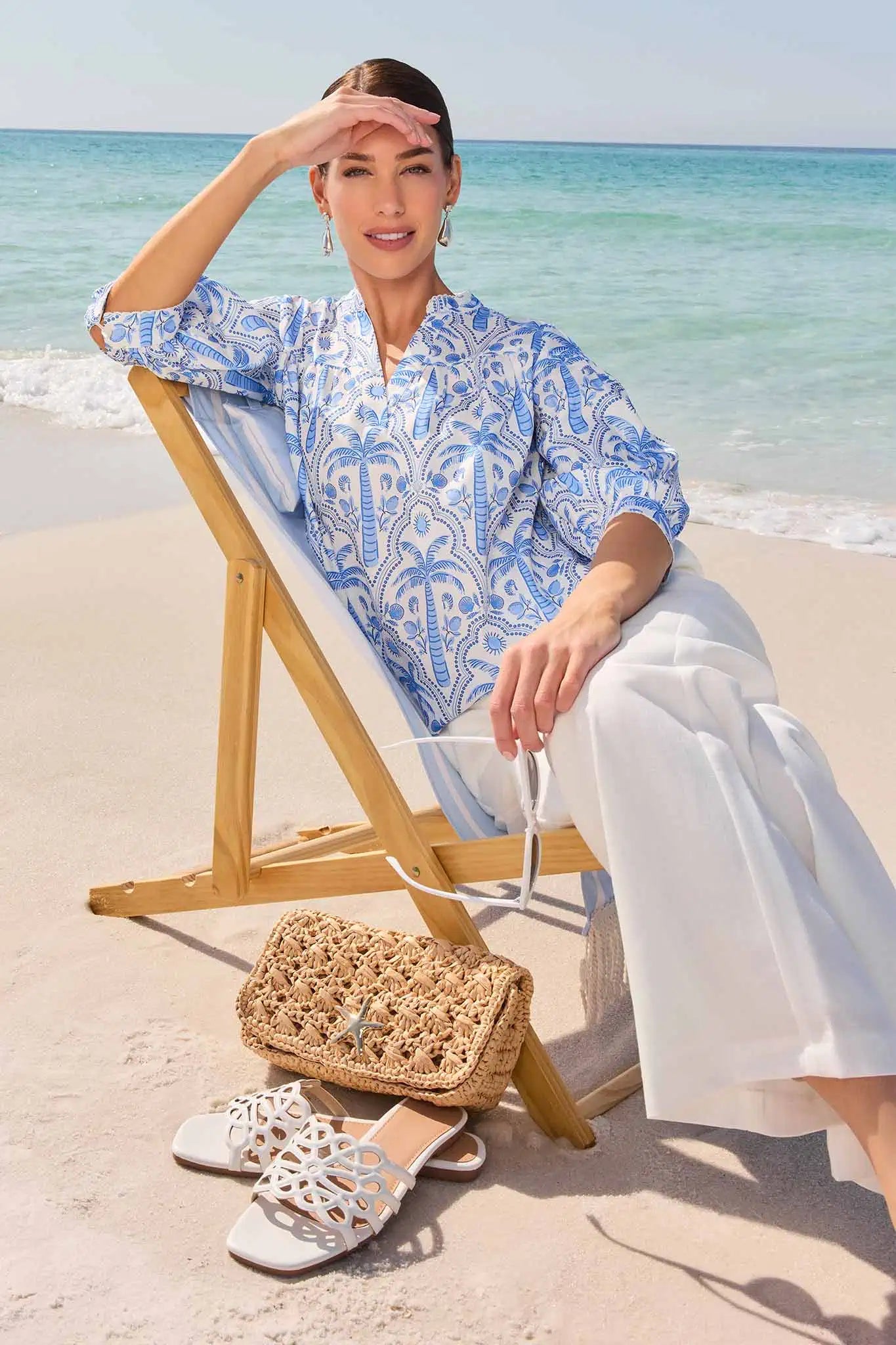 Woman in a blue patterned shirt and white pants sitting on a beach chair with a woven bag and sandals beside her.