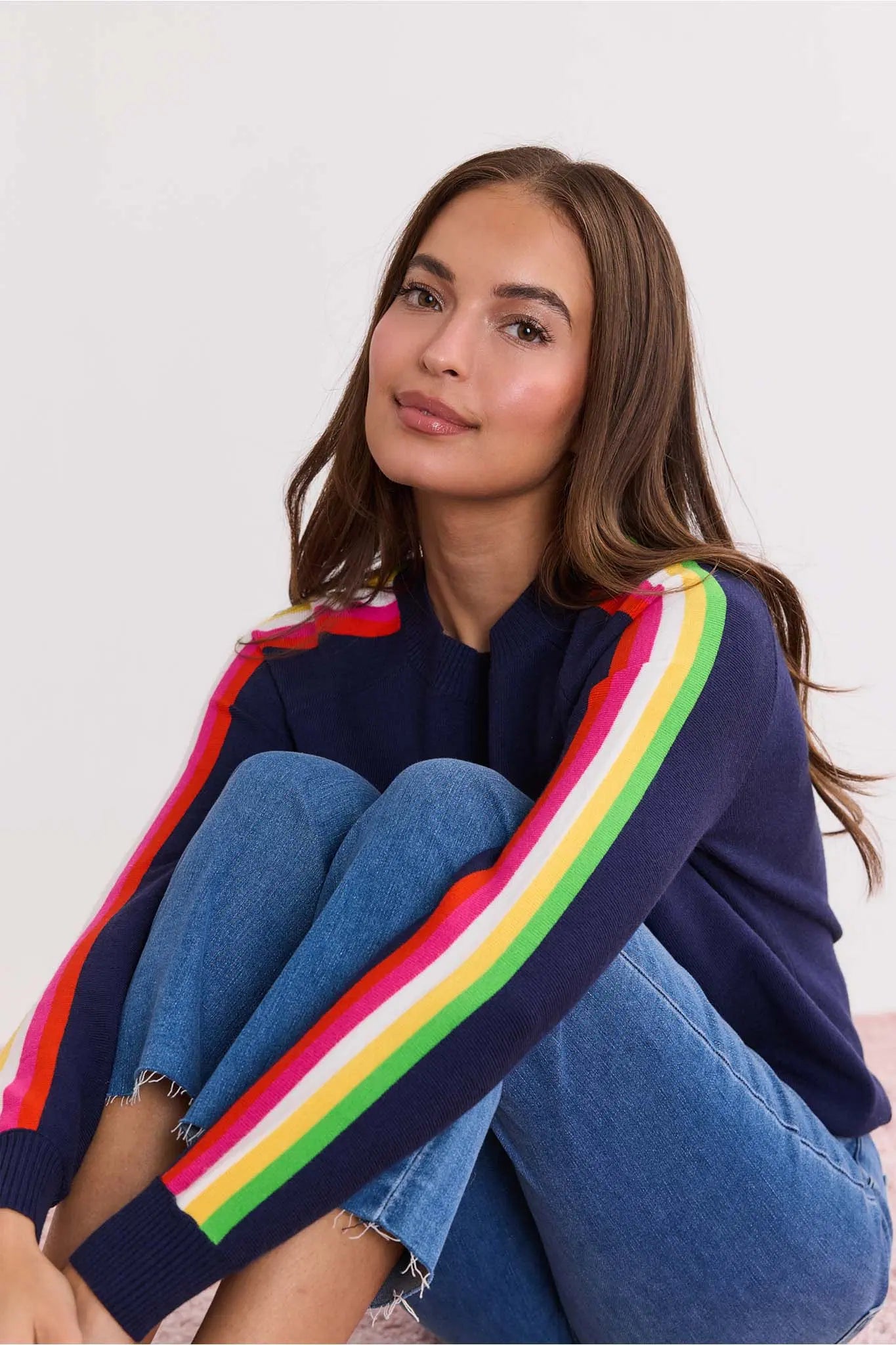 Woman wearing a Khloe Navy Striped Sleeve Sweater with colorful stripes sitting on the floor against a white background
