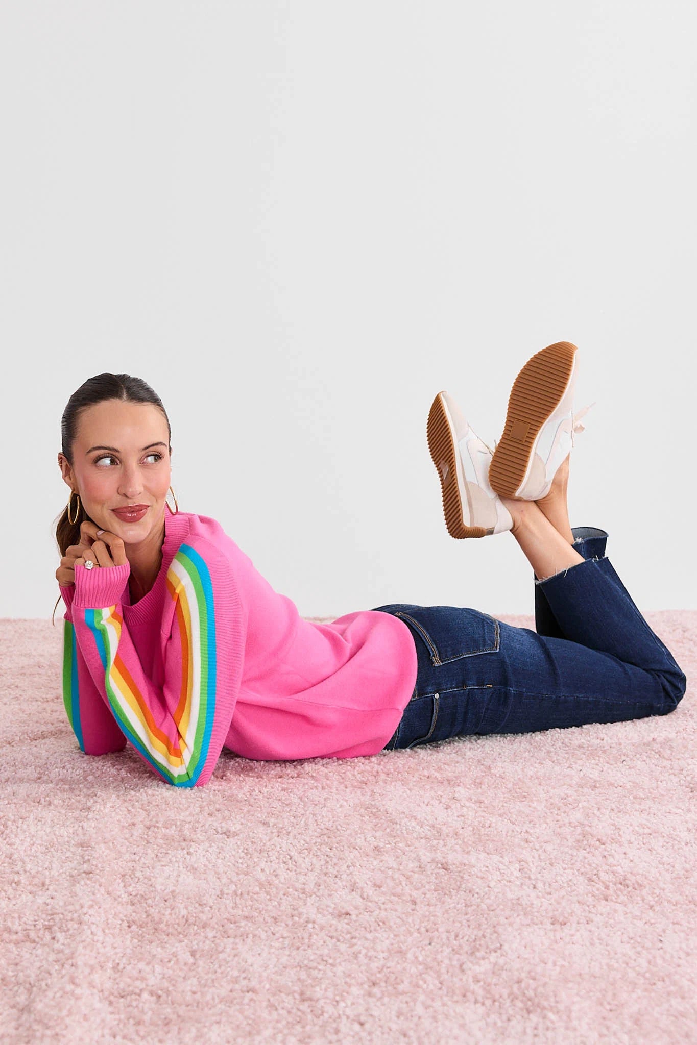 Woman wearing a Khloe Pink Striped Sleeve Sweater with a rainbow design and blue jeans, lying on a pink carpet.