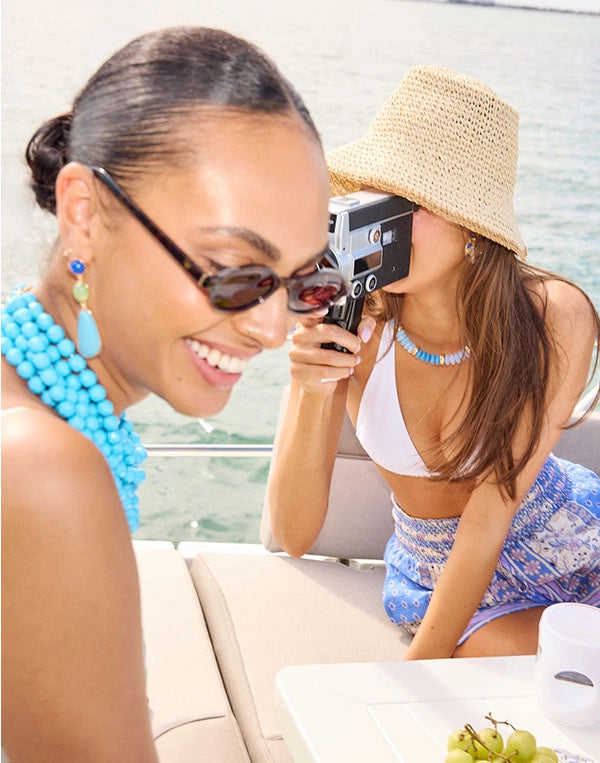 Two women on a boat, one taking a photo with a camera, wearing sunglasses and a blue beaded necklace.