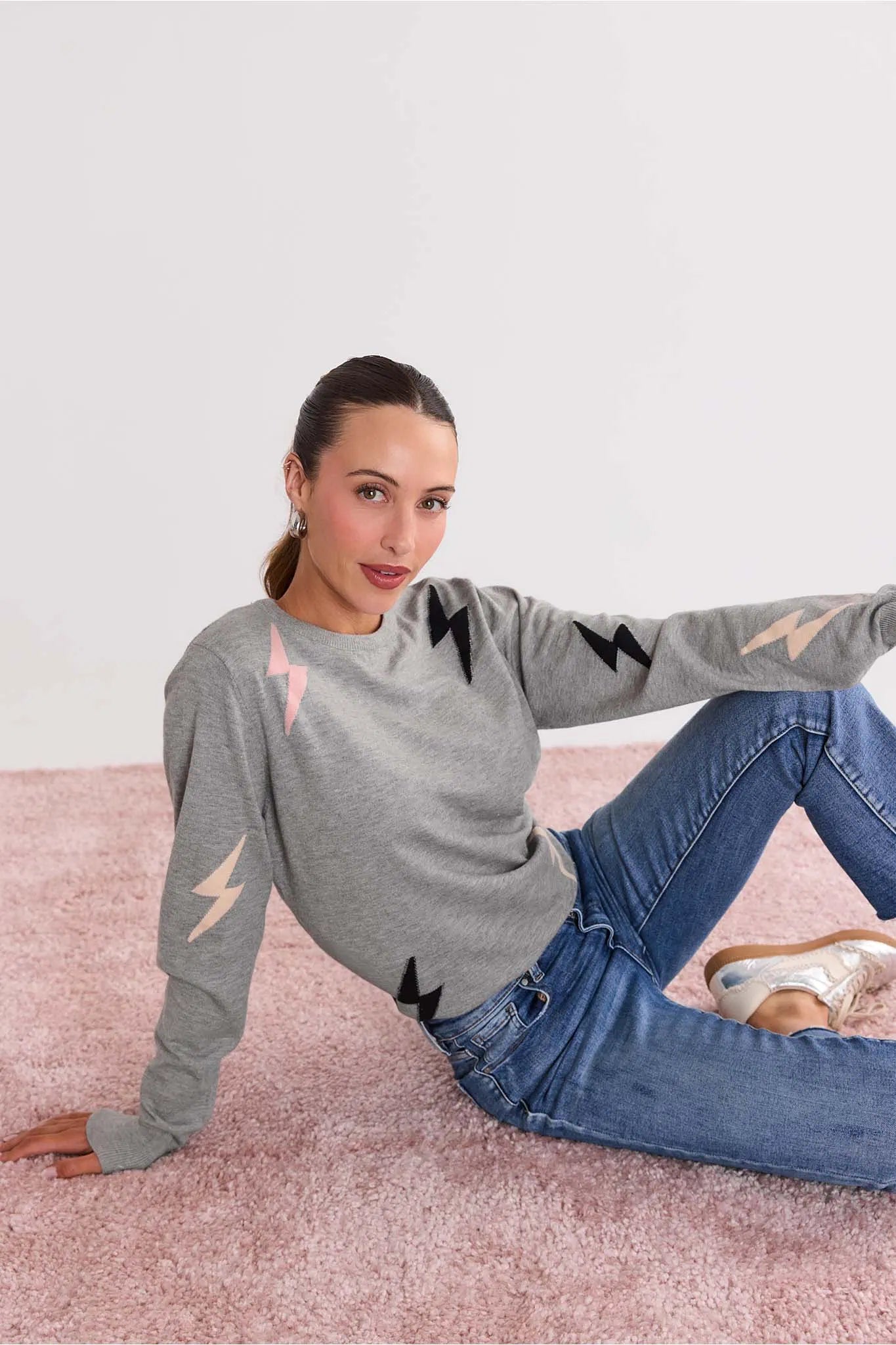 Woman wearing a Lightning Bolt Gray Knit Sweater and blue jeans sitting on a pink carpet.